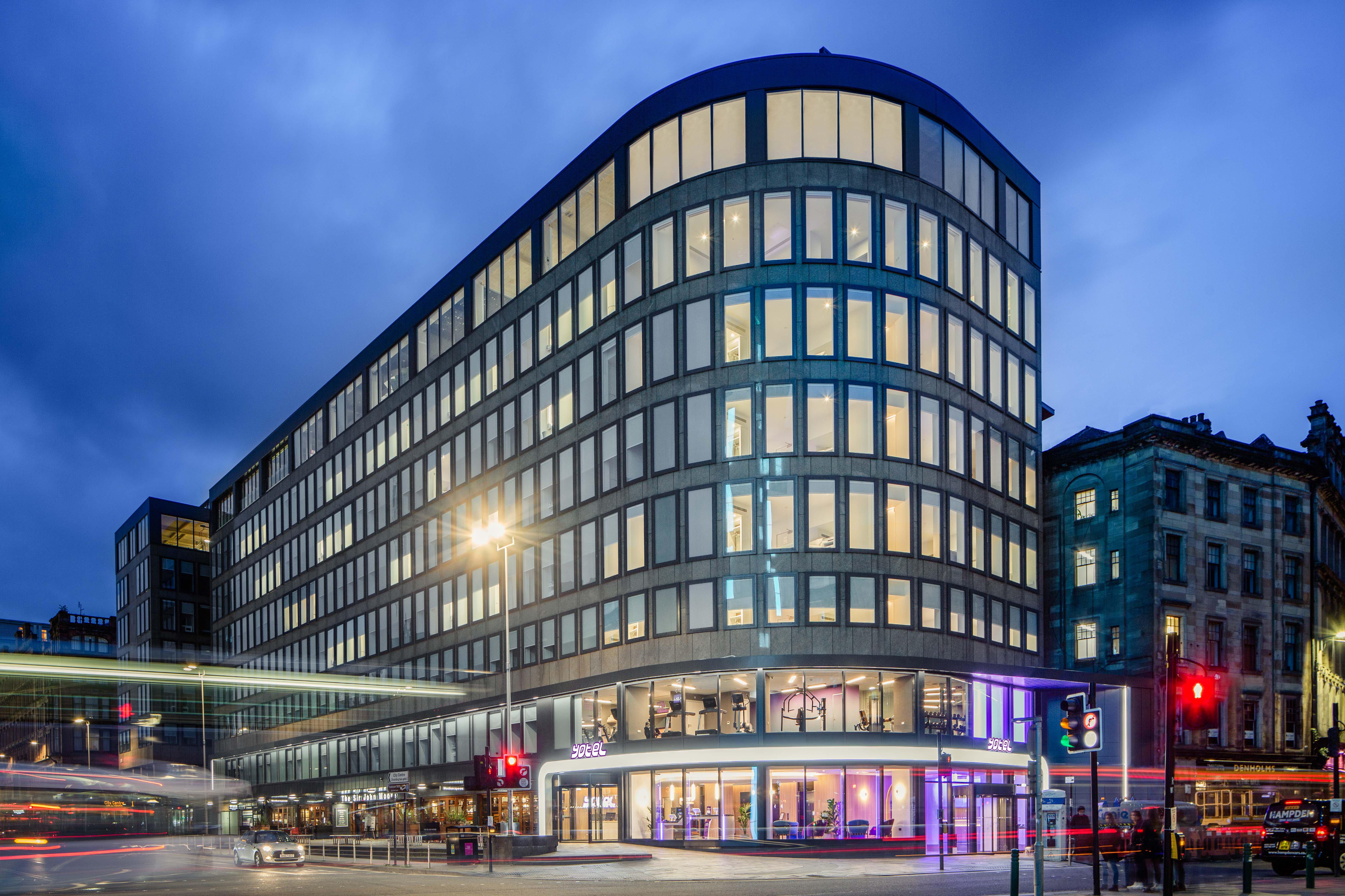 Yotel Glasgow Ngoại thất bức ảnh The photo shows a modern office building with a distinctive curved shape and a facade made of large glass windows. It is illuminated against a twilight sky, giving it a contemporary appearance. The ground floor features large glass entrance areas, li